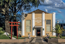 Imagem: Fotografia C: Prédio amarelo com colunas brancas pela estrutura. Na frente há bancos e árvores em área de vegetação. O prédio indica “biblioteca Publica Municipal, 1958” Fim da imagem.