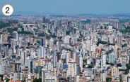 Imagem: Fotografia. 2: Vista aérea de prédios altos cobrindo grande parte da cidade até o horizonte da foto. Fim da imagem.