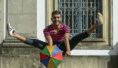 Imagem: Fotografia. Homem de cabelo curto castanho, sorrindo, vestindo camiseta rosa com listras azuis e calça preta.  Fim da imagem.