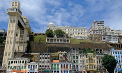 Imagem: Fotografia. Vista de casas antigas na margem de um morro. Acima do morro há casarões e monumento retangular com passarela para vista do mar. Fim da imagem.