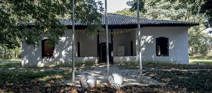 Imagem: Fotografia. Vista de casa branca com porta central de arco e duas janelas pequenas em arcos laterais. No centro há uma pequena varanda. Ao redor há árvores e gramado. Fim da imagem.