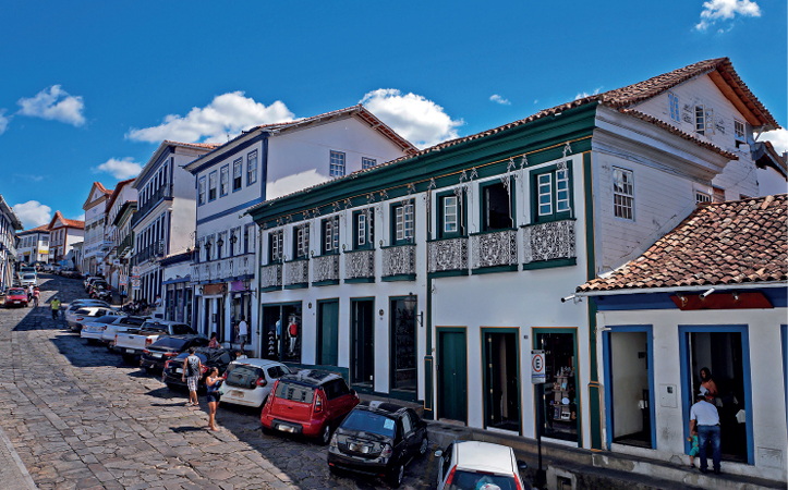 Imagem: Fotografia. Vista de rua com casas antigas de dois e três andares em rua de ladeira. Todas brancas com janelas e portas coloridas com abertura direto para calçada. Fim da imagem.