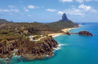 Imagem: Fotografia. Vista aérea de morros de pedra e vegetação baixa à margem de uma praia com mar azul cristalino. É possível ver a parte submersa de rochas próximas a praia. Fim da imagem.