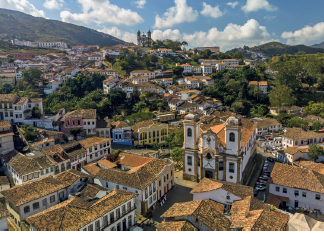 Imagem: Fotografia. Vista aérea de cidade construída em morros. Há casarões e igreja antiga. Entre as construções há vegetação.  Fim da imagem.