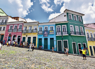 Imagem: Fotografia. Vista de ladeira de pedra com casas coloridas de dois e três andares lado a lado. As portas abrem diretamente na calçada. Há pessoas caminhando na rua. Fim da imagem.