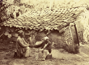 Imagem: Fotografia. Casa baixa de madeira e ferro com telhado irregular. À frente, homem e mulher idosos sentados em banquinhos de pedra. Fim da imagem.