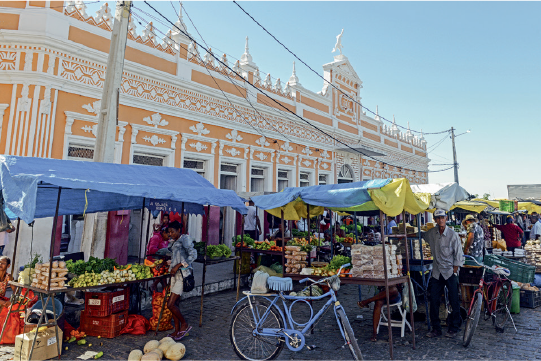 Imagem: Fotografia. Vista de rua com barracas de frutas e verduras expostas. Há pessoas transitando.  Fim da imagem.