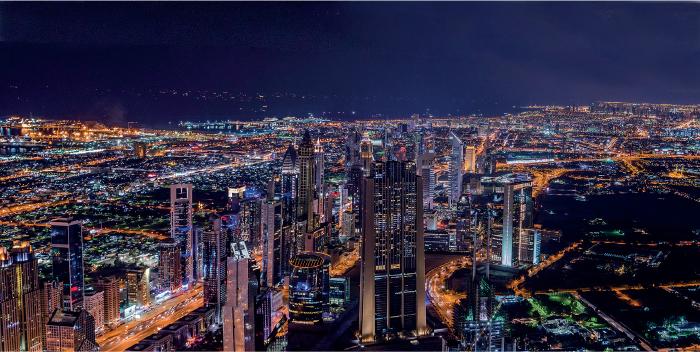 Imagem: Fotografia. Vista aérea de extensão longa de uma cidade iluminada a noite por luz elétrica das casas, prédios e ruas. Fim da imagem.