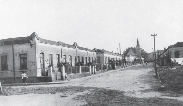 Imagem: Fotografia em preto e branco. Vista de rua de terra com casas de muro e grade baixa. Ao fundo, uma igreja com árvores. No canto direito da rua há postes de madeira. Fim da imagem.