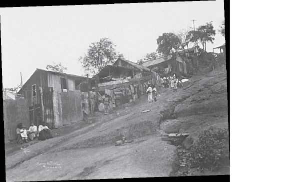 Imagem: Fotografia em preto e branco. Vista de morro com casas antigas de madeira e pessoas na frente.  Fim da imagem.