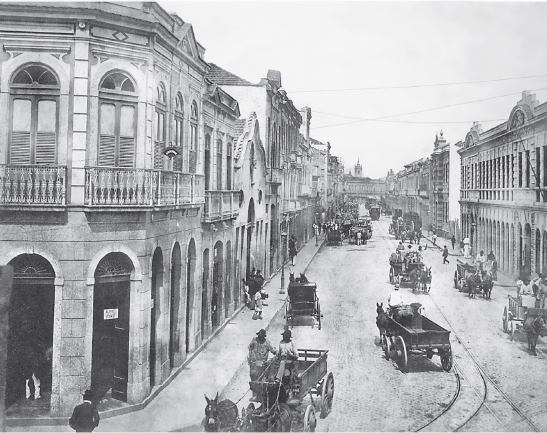 Imagem: Ilustração em preto e branco. Rua com casarões altos de janelas e portas compridas. Sobre a rua larga de terra há carroças puxadas por cavalos e pessoas transitando.  Fim da imagem.