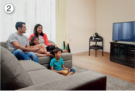 Imagem: 2. Fotografia. Família sentada em um sofá largo cinza, estão em frente a uma televisão retangular fina sobre um móvel preto em uma sala. Fim da imagem.