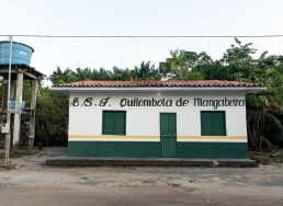 Imagem: Fotografia. Faixada de casa branca pequena com porta central e janela nas laterais verdes. Acima, o letreiro diz “E.S.F. Quilombola de Mangabeira”. Ao redor há palmeiras e árvores, ao lado há uma caixa de água. Fim da imagem.