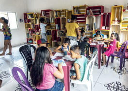 Imagem: Fotografia. Interior de uma sala com crianças sentadas em mesas e próximas a estantes com livros diversos. Fim da imagem.