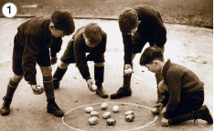 Imagem: Fotografia em preto e branco. 1: Cinco meninos de cabelo curto, vestindo casaco e bermuda. Estão jogando bolinhas em círculo pintado sobre o chão.  Fim da imagem.
