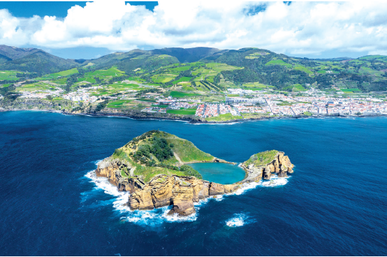 Imagem: Fotografia. Vista aérea de pequena ilha formada por rochas com vegetação ao redor, e lago no centro. Ao fundo ilha extensa com vegetação e casas na beira do mar. Fim da imagem.