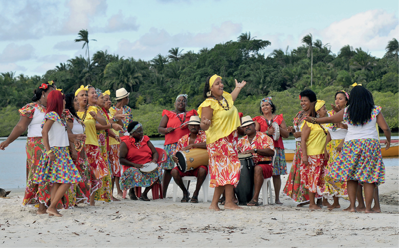 Imagem: Fotografia complementar das páginas 76 e 77. Mulheres de roupas coloridas em tons de amarelo, branco e vermelho. Estão cantando juntas sobre a praia. Há homens e mulheres sentados entre elas tocando instrumentos musicais. Ao fundo, vegetação fechada. Fim da imagem.