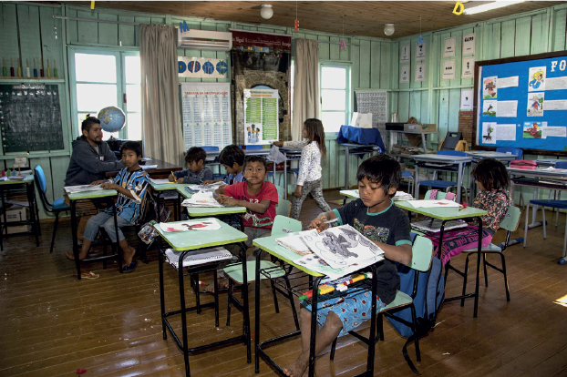 Imagem: Fotografia. Crianças indígenas em uma sala de aula sentados em carteiras individuais. Fim da imagem.