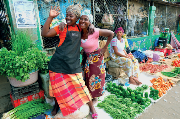 Imagem: Fotografia. Mulheres de turbante colorido, vestindo vestido e avental. Estão em frente a uma banca com verduras dispostas em bancada e bandejas. Fim da imagem.