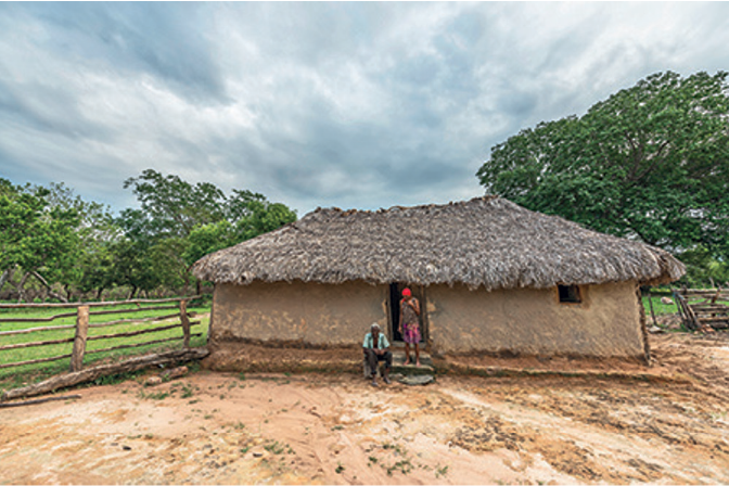 Imagem: Fotografia. Vista de casa de barro retangular com telhado de palha. Há uma mulher sobre a frente da casa, de touca vermelha, camiseta marrom e saia rosa, e um homem de cabelo curto grisalho, vestindo camisa verde e calça preta, sentado em um banco de madeira. À frente da casa há chão de terra e atrás há árvores e gramado. Fim da imagem.