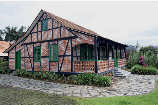 Imagem: Fotografia. Casa de tijolo a vista com telhado triangular longo indo do inicio ao fim da casa baixa. Ao redor há caminho de pedras e flores. Fim da imagem.