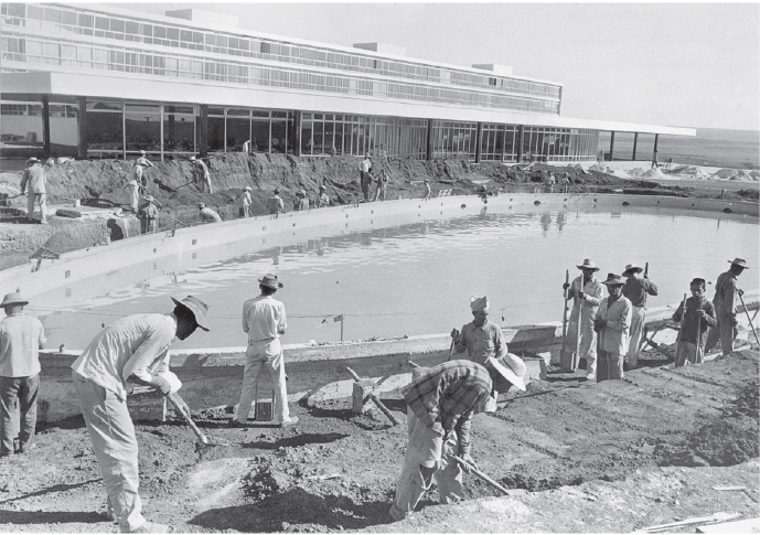 Imagem: Fotografia em preto e branco. Homens ao redor de uma fonte em construção. Atrás há um prédio em construção. Fim da imagem.