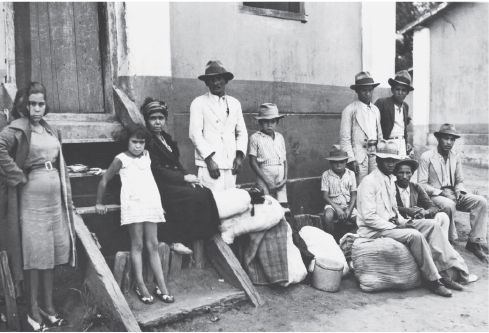 Imagem: Fotografia em preto e branco. Homens, mulheres e crianças sentados em frente a uma casa de alvenaria com porta e escada de madeira.  Fim da imagem.