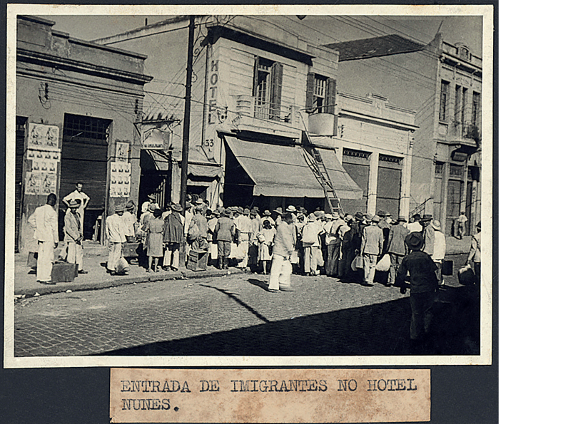 Imagem: Fotografia em preto e branco. Aglomerado de pessoas em frente a um hotel com toldo aberto sobre a calçada. Pessoas carregam malas e sacos. Fim da imagem.