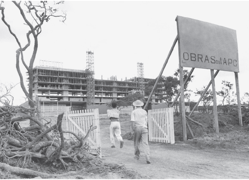 Imagem: Fotografia. Pessoas em estrada de terra entrando por um portão de madeira. Ao fundo há a estrutura de um prédio em construção. Fim da imagem.