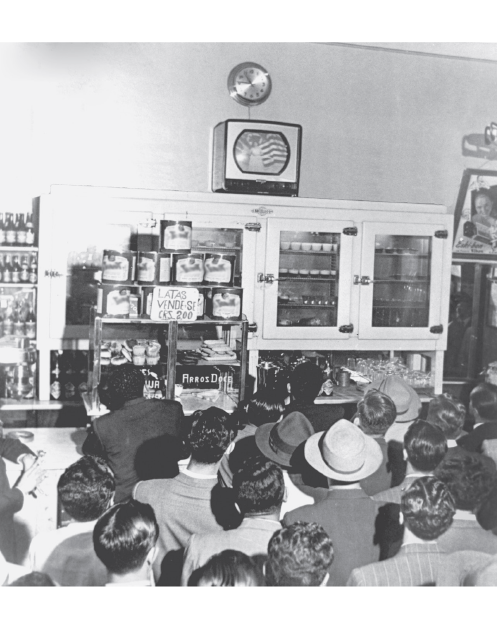 Imagem: Fotografia em preto e branco. Homens em uma sala fechada vendo uma televisão quadrada de tubo em cima de uma estante. Fim da imagem.