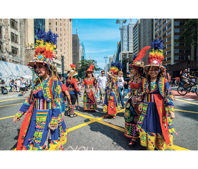 Imagem: Fotografia. Mulheres vestindo vestido longo colorido e chapéu com plumas. Estão andando em uma rua extensa com prédios ao redor. Fim da imagem.