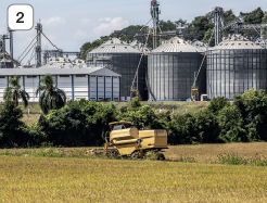 Imagem: Fotografia. 2: Campo aberto com um trator passando pela plantação. Ao fundo, há tanques grandes cinzas ligados a usina.  Fim da imagem.