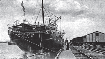 Imagem: Fotografia em preto e branco. Navio grande com vela içada atracado sobre porto. Ao lado, há um trilho e pessoas entre a calçada. Fim da imagem.