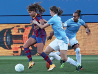Imagem: Fotografia. Três mulheres jogando bola em um campo de futebol. Uma delas possui cabelo longo castanho, vestindo camiseta vermelha e azul, as outras duas possuem cabelo preto, vestindo camiseta azul clara. Fim da imagem.