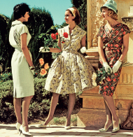 Imagem: Fotografia. Fotografia. Três mulheres de vestido curto, segurando flores em frente a um muro com alto relevo desenhado. Mulher de cabelo curto preto, vestindo vestido liso verde claro. Mulher de cabelo médio castanho e faixa amarela, vestindo vestido de flores estampadas brancas. Mulher de cabelo médio ruivo e chapéu verde, vestindo vestido curto com flores marrom e vermelha. Fim da imagem.