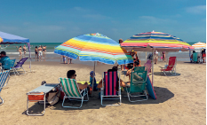 Imagem: Fotografia. Vista de pessoas sentadas em cadeiras sobre areia da praia. Ao fundo há pessoas sobre o mar. Fim da imagem.