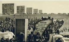 Imagem: Fotografia em preto e branco. Há multidão de pessoas reunidas em campo aberto com prédios e carros ao redor. Em destaque, um cartaz em forma de mapa do brasil que diz “Juscelino – Acordou o Gigante Adormecido, Brasília – A capital da esperança, Salve JK.”. Fim da imagem.