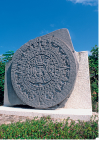 Imagem: Fotografia. Escultura de pedra arredondada com ornamentos em alto relevo formando a imagem de um rosto no centro com formas formando padrões ao redor.  Fim da imagem.