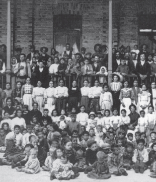 Imagem: Fotografia em preto e branco. Crianças reunidas em frente a um pátio, sentadas no chão. Atrás há homens e mulheres em pé. Todos estão pousando para fotografia de mateira ereta com braços lado a lado. Fim da imagem.