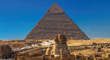 Imagem: Fotografia. Esfinge a frente de uma pirâmide. Monumentos sobre o deserto com runas aos arredores. Fim da imagem.