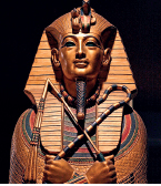 Imagem: Fotografia. Túmulo egípcio em forma de faraó dourado com tons de azul. Ele possui ornamentos na cabeça, pescoço e queixo. Está com os braços cruzados, segurando um chicote e um cajado. Fim da imagem.