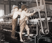 Imagem: Fotografia. Crianças descalças trabalhando em cima de maquinário com fios entrelaçados entre engrenagens da máquina. Fim da imagem.