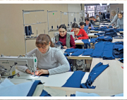 Imagem: Fotografia. Mulheres trabalhando em linha de produção de tecidos com mesas enfileiradas com máquina de costura. Fim da imagem.