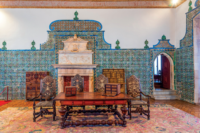 Imagem: Fotografia. Vista de sala aberta com mesa de madeira e cadeiras com estampas ornamentadas. Ao redor, azulejos azuis com desenhos de arabescos. No centro, uma lareira. Fim da imagem.