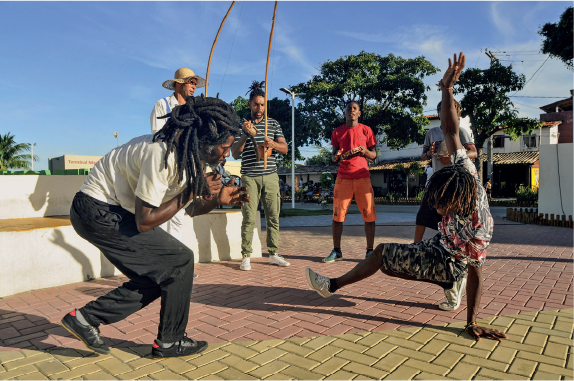 Imagem: Fotografia. Homens em uma praça jogando capoeira. Atrás de dupla duelando há três homens tocando instrumentos musicais. Fim da imagem.