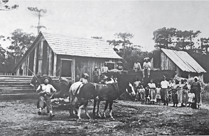 Imagem: Fotografia em preto e branco. Vista de casas de madeira com pessoas sentadas sobre pátio de terra posando para fotografia. Há cavalos entre as pessoas. Fim da imagem.