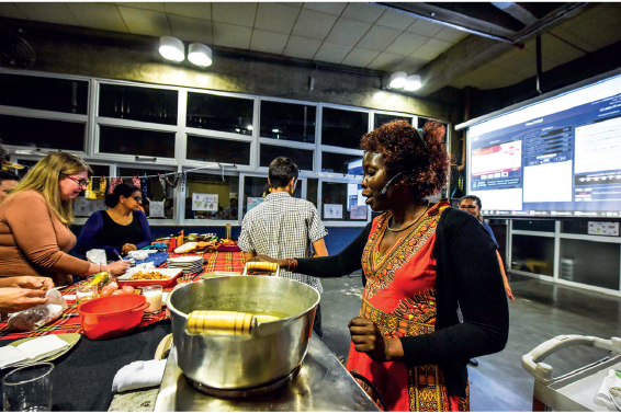 Imagem: Fotografia. Destaque de mulher de cabelo cacheado castanho, vestindo vestido vermelho e casaco azul. Está em frente a bancada com panelas e potes com comida. Atrás da mulher há um telão ligado com informações e imagens. Fim da imagem.