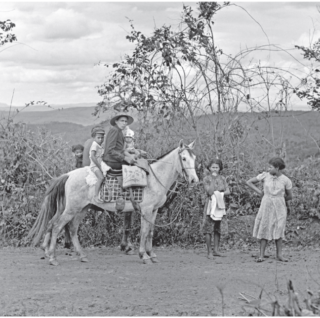 Imagem: Fotografia em preto e branco. Homem montado em cavalo com três crianças na garupa. À frente, menina e mulher. Estão todos em uma estrada de terra com vegetação seca ao redor. Fim da imagem.