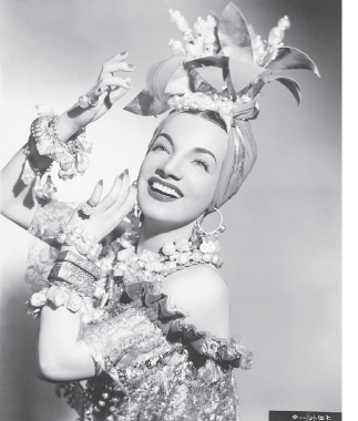 Imagem: Fotografia em preto e branco. Mulher de turbante com formas de frutas penduradas na cabeça. Veste um vestido com estampa e babado na altura dos ombros. Fim da imagem.
