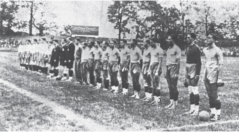 Imagem: Fotografia em preto e branco. Fileira de homens lado a lado sobre um campo de futebol. Eles estão uniformizados com camiseta e shorts. Fim da imagem.
